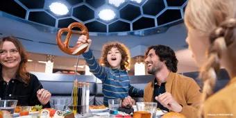 A child holding up a giant pretzel while sat at a table eating food with his two parents and sibling