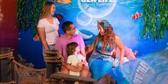 SEA LIFE Orlando Aquarium Mermaid with blue tail and seashells in her hair meeting a family including parents and a small kid. In the background there is a SEA LIFE sign.