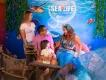 SEA LIFE Orlando Aquarium Mermaid with blue tail and seashells in her hair meeting a family including parents and a small kid. In the background there is a SEA LIFE sign.