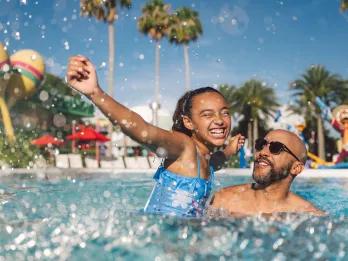 Disney's All-Star Music Resort Dad lifting daughter up in the resort pool, both smiling. There are giant statues of musical instruments.