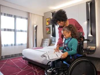 Disney Hotel New York – The Art of Marvel Hotel room with burgundy carpet and double bed and a girl in a wheelchair reading a book and her mum stood over her.
