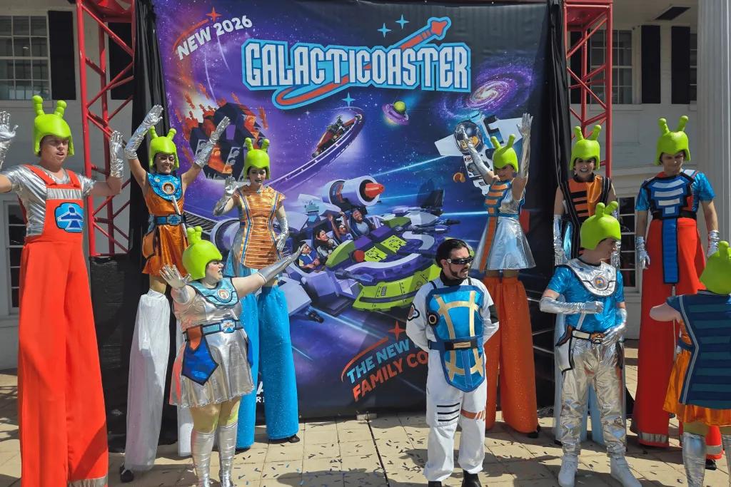 The Galacticoaster People with alien green helmets on and alien outfits, some stood on stilts, stood in front of a ride poster.