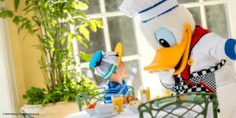 Donald Duck dressed as a chef leaning down to greet a child sat at a table wearing a Donald Duck hat
