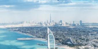 View from over the crystal clear sea with a view of the beach with an island off of it with a sail shaped building on it called the Burj Al Arab. In the background you can see a view of the city skyline and the Burj Khalifa.