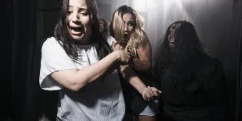 Two young women in a dark room with a bright light shining behind with terrified facial expressions, clutching onto one another and dodging another person who is lurking in the dark. The third person has black hair and a wound on their head which seems to be bleeding down their face. Their hair is messy and they are pulling a scary face with their tongue poking out.
