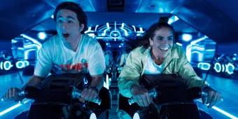 Two young adults sat on motorcycles shaped vehicles TRON Lightcycle. They are leaning forward gasping with shock. 