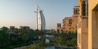 Beige buildings with a river beside them and some trees. It is sunset and the view shows the Burj Al Arab, a ship sail shaped building.