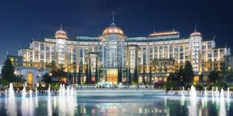 Large white building with rounded shape around large fountains. On top there are circular details on the roof that are golden with turquoise spikes on top. Looks very modern.