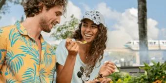 Girl smiling holding a plastic fork with food on it looking at a man who is holding the small food portion. In the background the monorail is driving past.