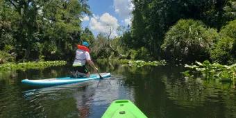 A person sat on a green kayak and a young person on paddle board in front kneeling by the water.