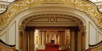 Golden and wooden bannisters lining two staircases which wrap around and join to create on large staircase. There is bright red carpet and white walls lined with gold details. There is a large open hallway with a fireplace at the end.