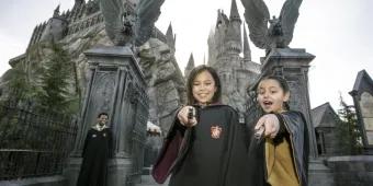 Two young girls wearing wizard robes and pointing magic wands at the camera while standing in front of Hogwarts Castle