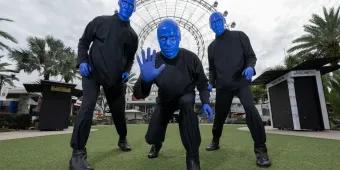 There are three men completely painted in blue paint stood in front of a big white Ferris wheel on a field. Each of them are posing in a strange way.