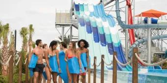 Group of six young adults holding blue mats walking away from large blue water slides. There are six identical slides with a small drop which leads to a bigger drop. 