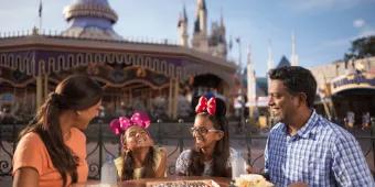 Family of four dining together in Magic Kingdom Park with carousel behind them. The two little girls are wearing minnie ears and smiling.