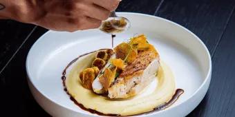 Michelin Dining in Orlando Chicken or fish dish on top of smooth mash potato with green plant on top and crackers. There is a mans hand pouting dark sauce around the dish on the clean white bowl.