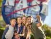 Kennedy Space Center Family of four, a man holding up a phone to take a selfie with wife and two kids, one boy and one girl. They are stood in front of a large sign, shaped like the earth, circular with red stripes over and the letters in white spelling NASA.