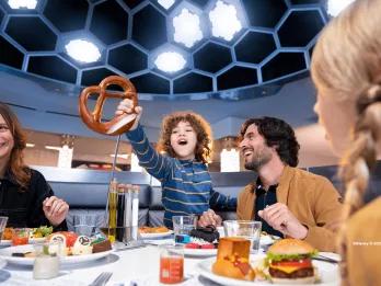 A child holding up a giant pretzel while sat at a table eating food with his two parents and sibling