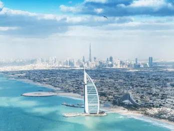 View from over the crystal clear sea with a view of the beach with an island off of it with a sail shaped building on it called the Burj Al Arab. In the background you can see a view of the city skyline and the Burj Khalifa.