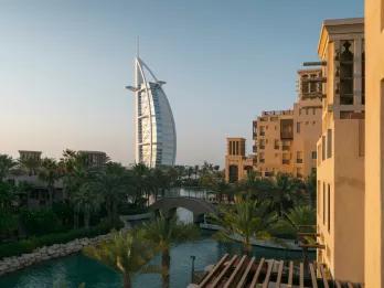 Beige buildings with a river beside them and some trees. It is sunset and the view shows the Burj Al Arab, a ship sail shaped building.