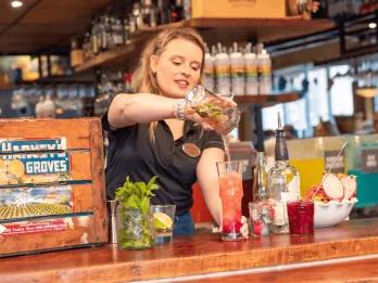 A woman pouring colourful drinks behind a wooden bar