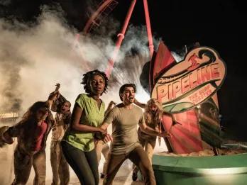 Group of young adults running through the park with a red rollercoaster in the background and fog. Through the fog you can see a scare actor with scary zombie makeup. 