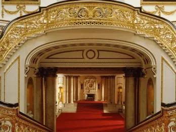 Golden and wooden bannisters lining two staircases which wrap around and join to create on large staircase. There is bright red carpet and white walls lined with gold details. There is a large open hallway with a fireplace at the end.