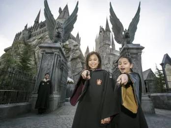 Two young girls wearing wizard robes and pointing magic wands at the camera while standing in front of Hogwarts Castle