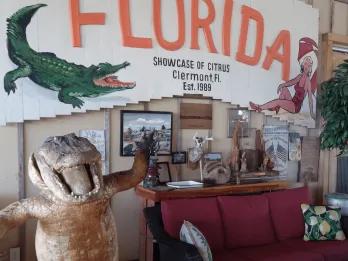 huge welcome to florida sign on the wall with image of a gator on and below there is a statue of an alligator stood up smiling and waving. Next to the statue there is an old sofa with old images above.