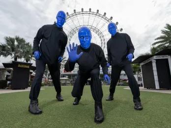 There are three men completely painted in blue paint stood in front of a big white Ferris wheel on a field. Each of them are posing in a strange way.