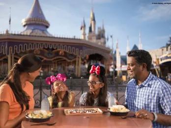 Family of four dining together in Magic Kingdom Park with carousel behind them. The two little girls are wearing minnie ears and smiling.