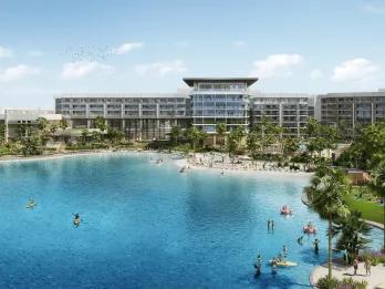 Large modern building which is rectangular stood behind a beautiful beach with sand and a huge pool shaped like the sea shore. There are people paddle boarding, kayaking and sitting on large inflatable flamingos.