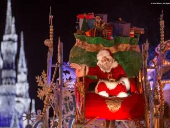 Santa Claus sat on a sleigh on a float full of presents. In the background Cinderella Castle is lit in white colours.