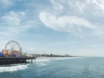 California There is the sea waves crashing onto a yellow sandy beach. There is also a long iconic pier with a funfair on it, including a Ferris wheel and small rollercoaster. In the far distance there are only a few large buildings..