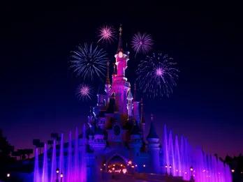 Castle lit neon pink in the night sky with purple fountains below. There are also pink fireworks in the sky.