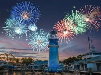 The iconic blue lighthouse at the front of the park with fireworks of many different colours exploding in the background above the silhouette of a ride,