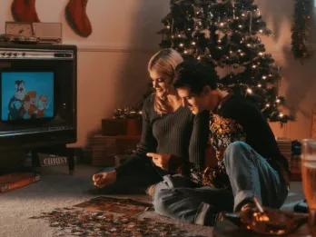 Couple sat playing with puzzle in front of a tv. Behind them there is a christmas tree and stockings hanging on the wall.