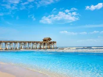 A long wooden pier with a shack at the end of it leading out to crystal clear water from a white sand beach.