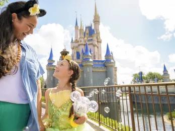 Magic Kingdom Park Mum holding the hand of her daughter who is dressed as Princess Tiana and holding a Mickey shaped bubble wand. In the background there's Cinderella Castle.